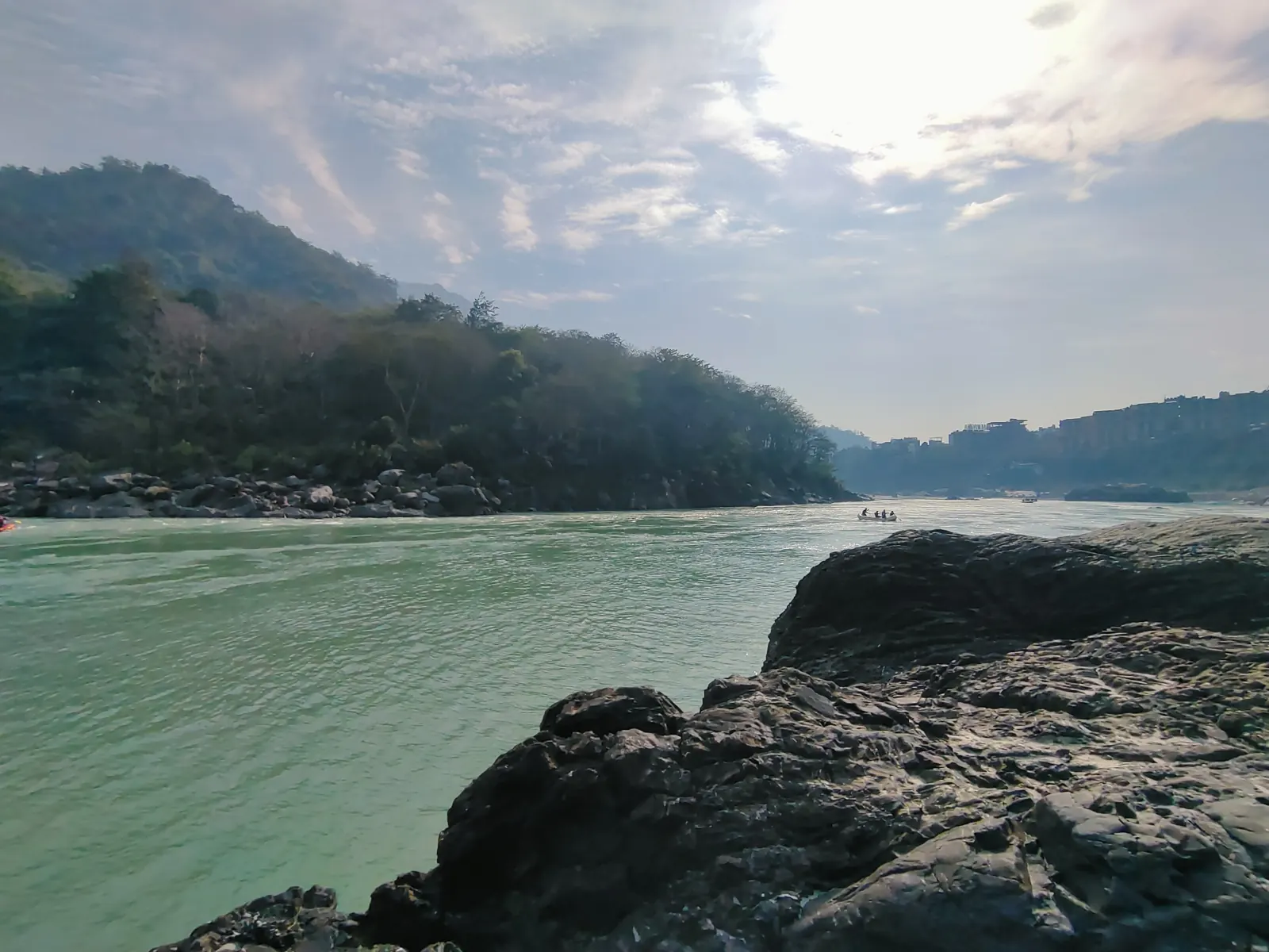 Jade-green Ganga flowing past large grey boulders below forested hills near Rishikesh